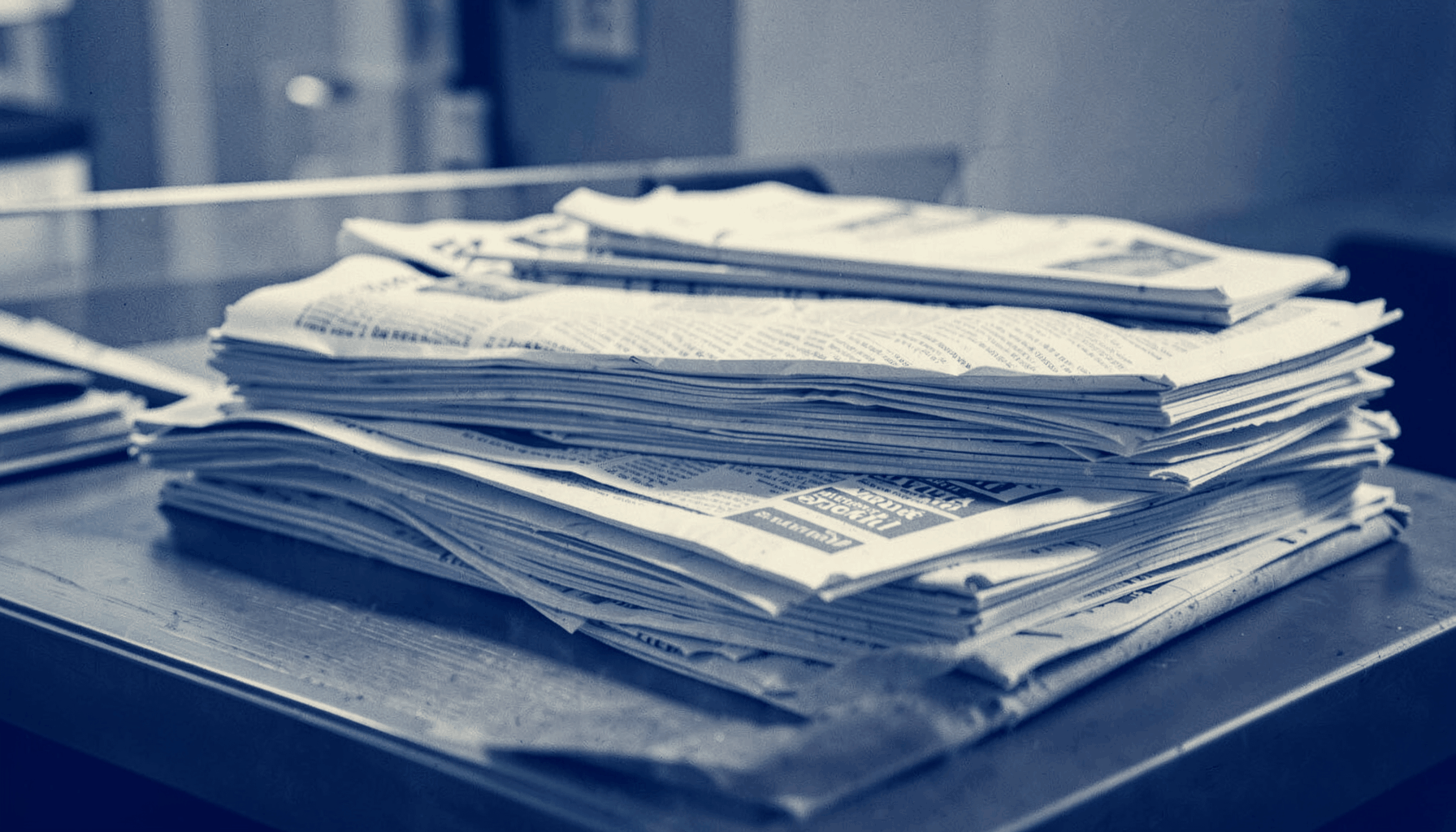 A duotone photograph in indigo and cream showing a neatly stacked collection of newspapers and magazines on a desk, with soft natural light falling across the pages.