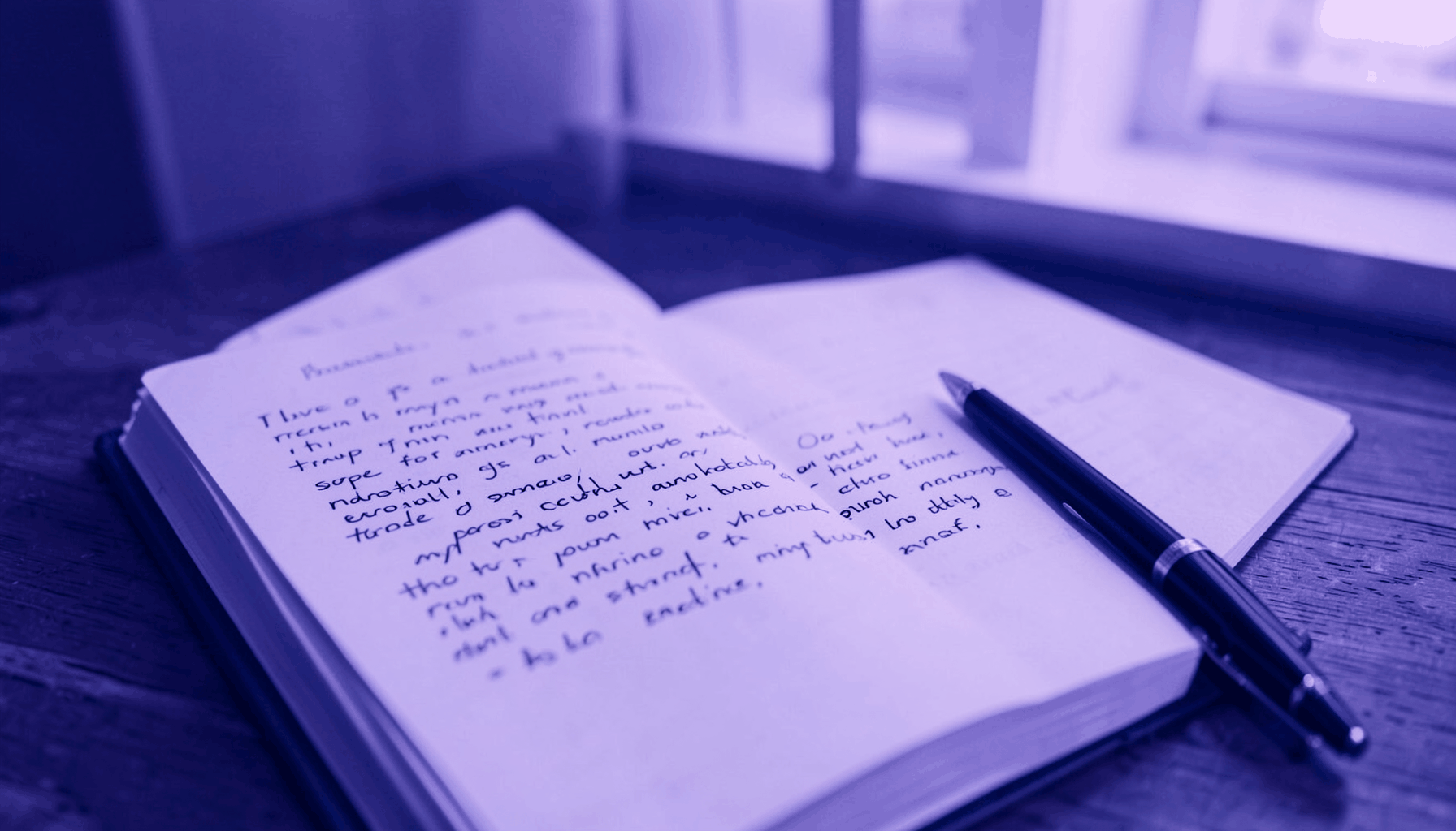 A duotone photograph in violet and navy showing an open notebook with handwritten text beside a pen, on a wooden desk with warm ambient light.